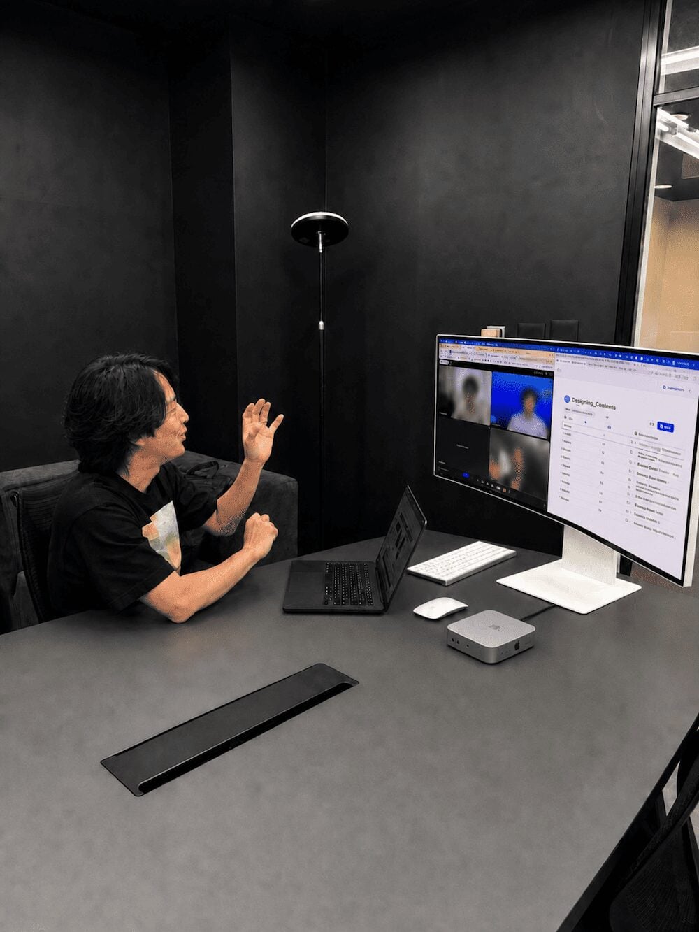 A person in a meeting room gesturing toward two monitors during a video call